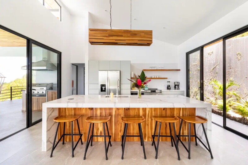 A kitchen island with five wooden stools and glass walls on either side looking out toward plants
