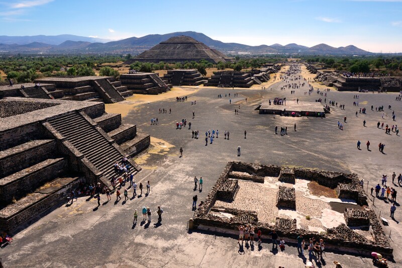 Aerial view of archaeological site, with one large stone pyramid in distance and many small ones, plus people walking among ruins