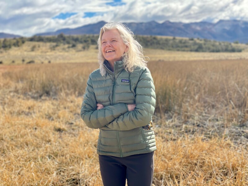 Woman wearing puffer jacket standing in a field and smiling with mountains in the background