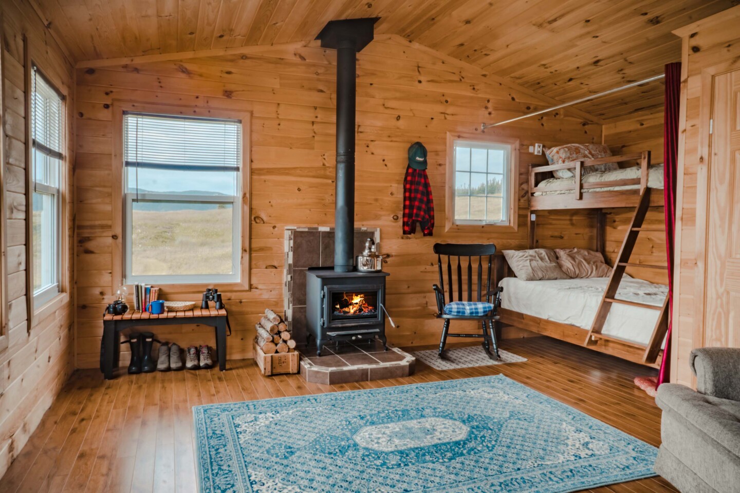 Interior of log cabin with several windows, black metal wood-burning fireplace, boots lined up against wall, and bunk beds