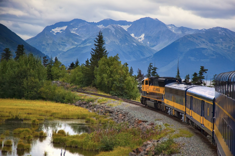 A blue-and-gold train passes a pond, with mountains in the background