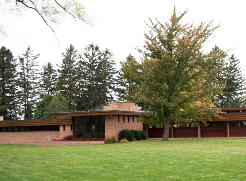 Green lawn in front of long brick and wood modernist house, with pine trees in distance
