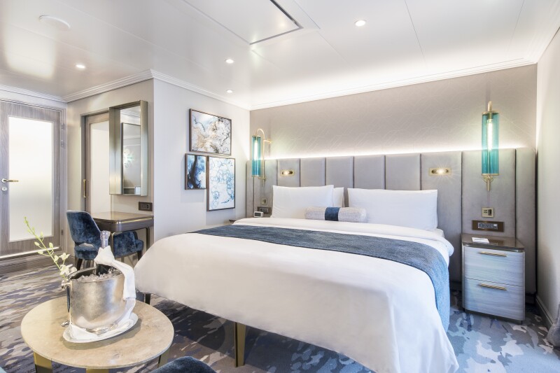 Image of a plush bed, desk and nightstands all with blue and gray accents in a veranda suite on a Crystal ocean ship