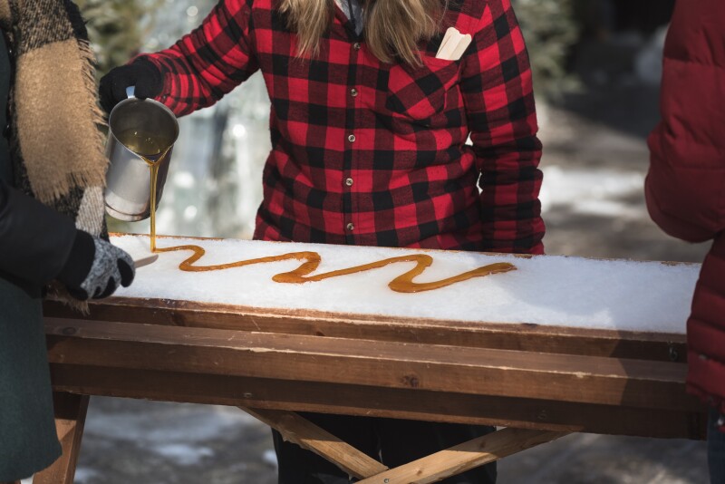 A person in a flannel shirt pouring a squiggle of maple syrup out of a metal pitcher onto a flat table of snow