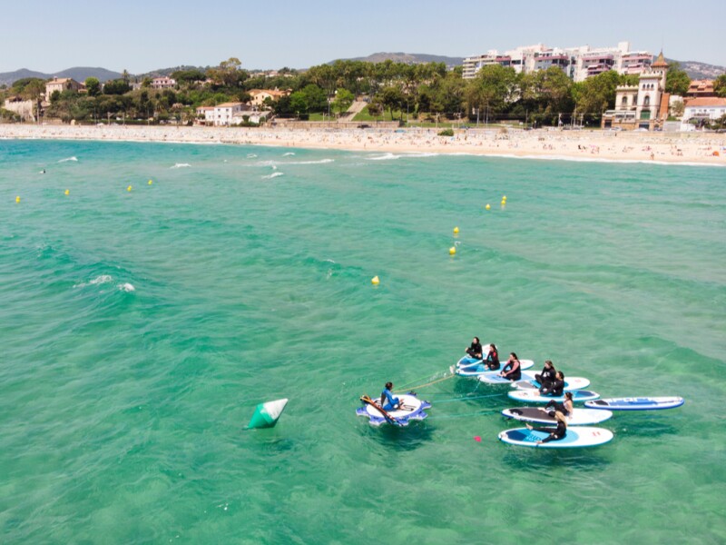 Half a dozen paddleboarders in green water off Playa de Ocata, with beach in distance