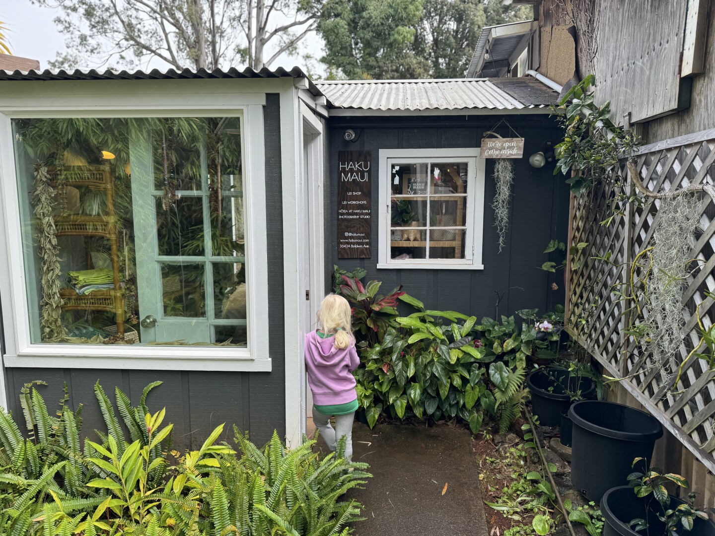 A young blonde girl leans against the door of a store named Haku Maui that's surrounded by foliage
