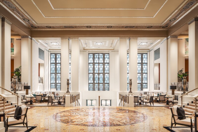 Spacious lobby, with high ceiling, large stained glass windows, stairs at left, right and center, and groups of sofas and armchairs in corners