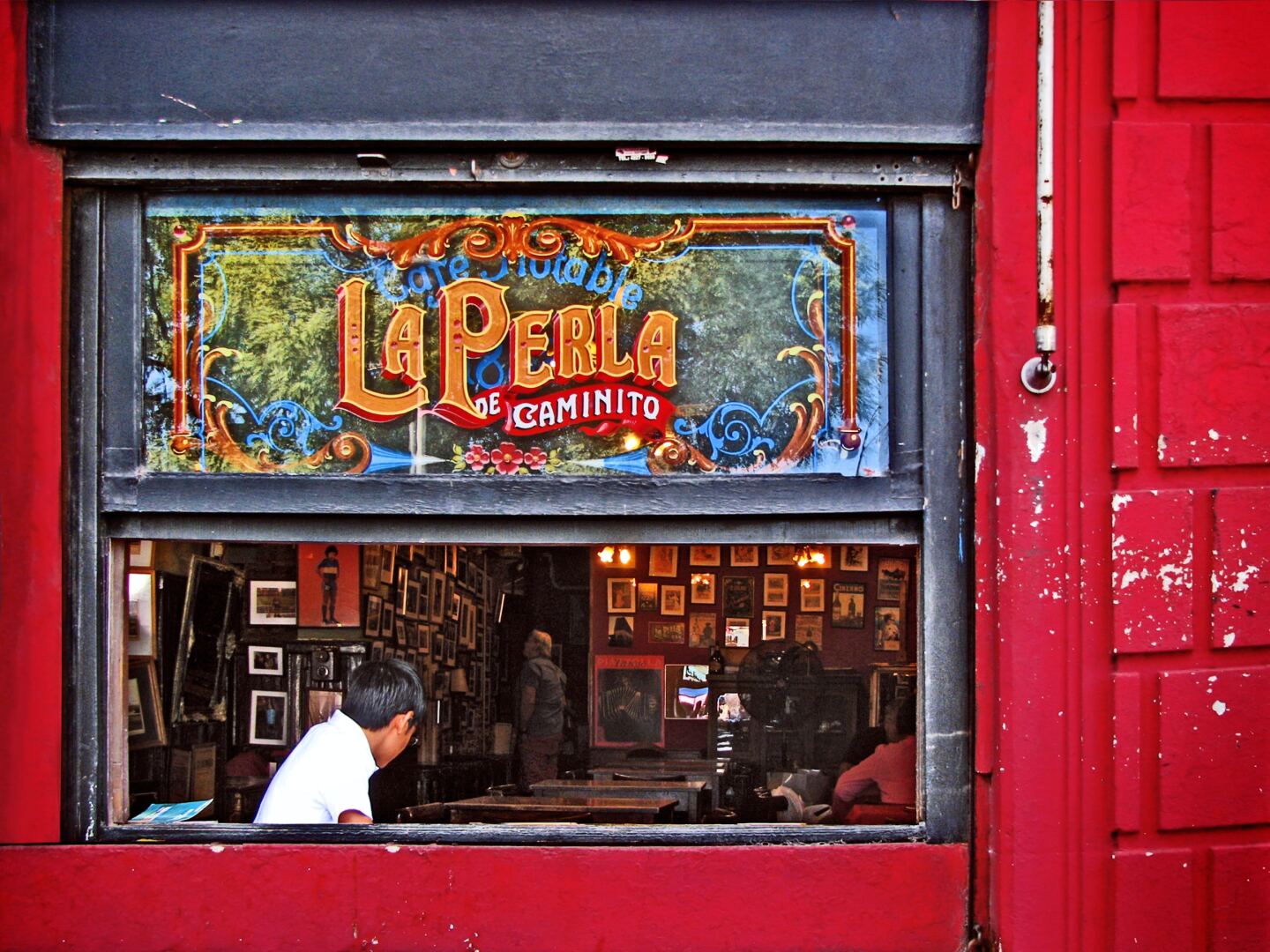 A bright red building with a large open window; inside is a man with a white shirt, under a sign for La Perla de Caminito café