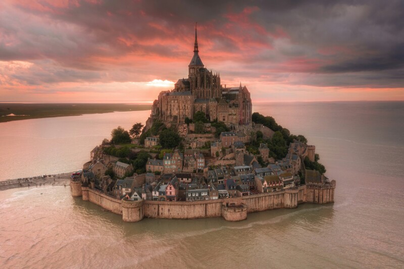 An aeriel view of an island commune topped with a striking abbey, in France, with moody sunset lighting and clouds