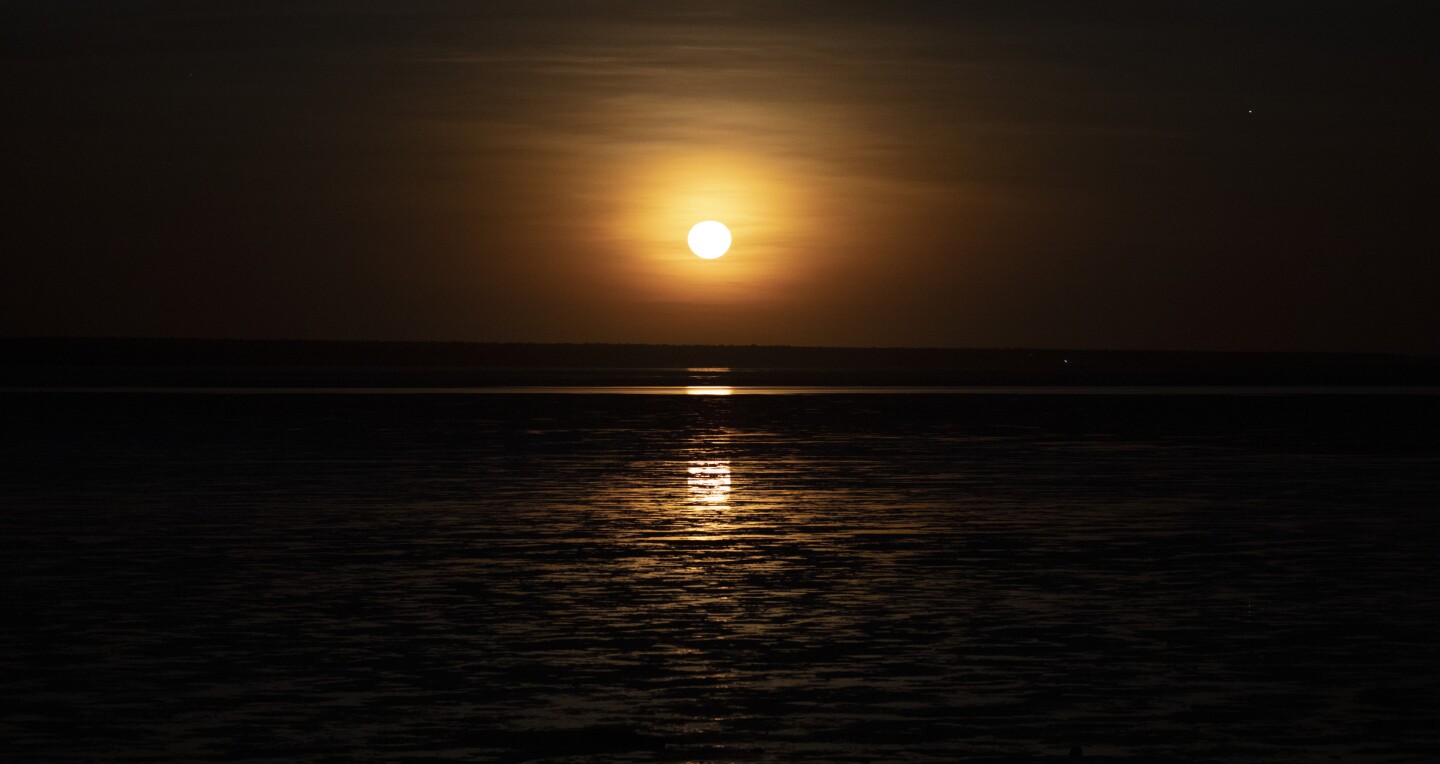 The full moon low in the sky over a bay glowing brightly with the Staircase to the Moon effect over Roebuck Bay, Broome, Australia.