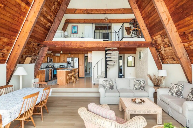 A multistory living and dining room of wood and white furnishings, with kitchen and spiral staircase in background