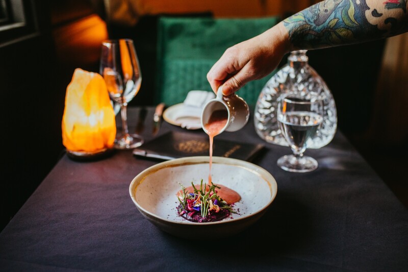 A tattooed arm pouring a reddish sauce over a plate of food, with glasses, decanter, and salt lamp in background