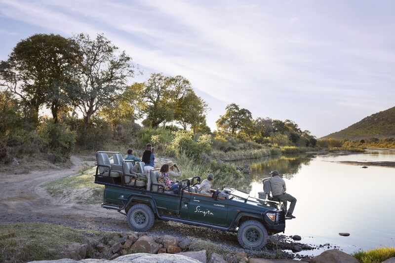 A family in roofless Jeep beside a small body of water while on a game drive at Singita Sabi Sand, with some trees along bank and a dirt road at left