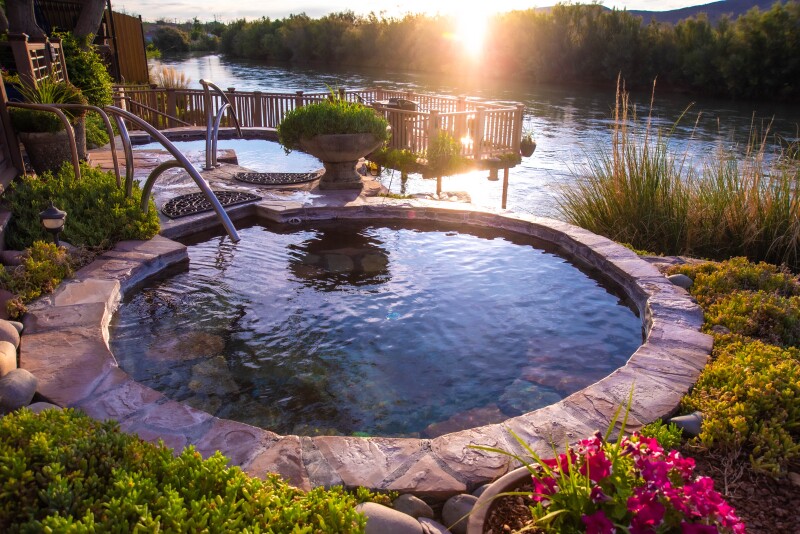 A large stone hot tub on landscaped grounds next to a river, with boardwalk over water in background