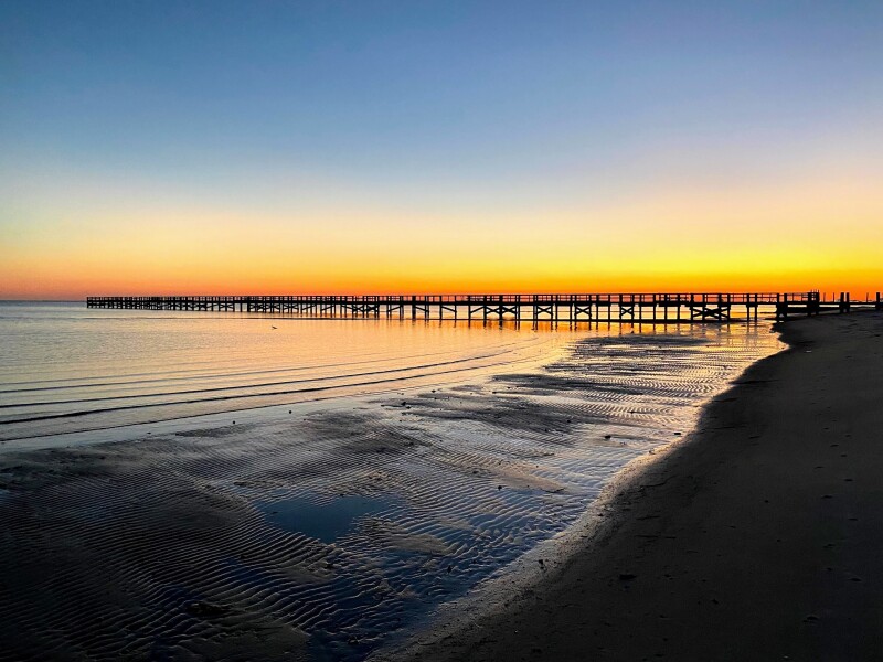 Sunset on dark beach, with a boardwalk jutting out into water in distance