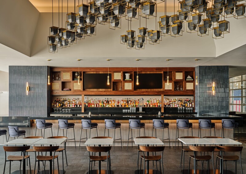 A fully stocked bar with wooden cabinetry above, blue tile details, hanging sconces and row of bar stools in background, with empty dining tables and chairs in foreground