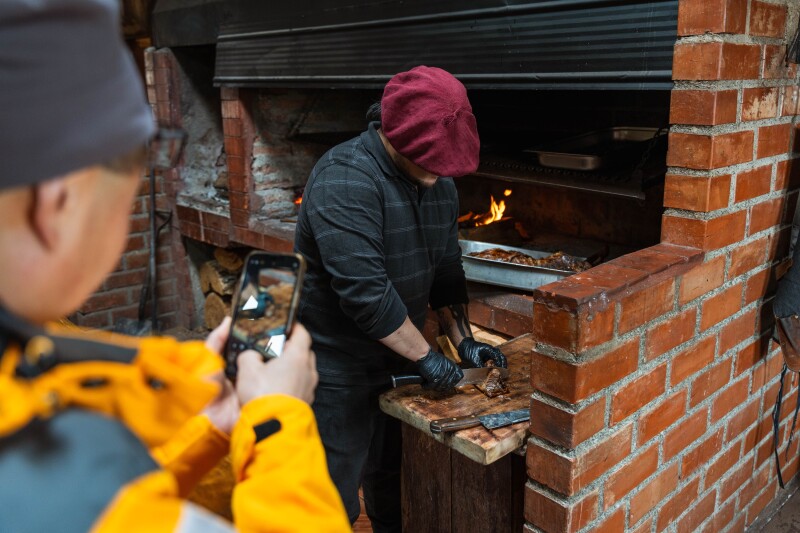 A person photographing on their phone a man who is chopping up meat that has just emerged from a wood fire grill