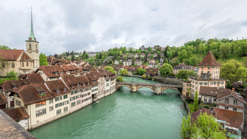 Historic buildings with brown roofs line a river crossed by a short, low bridge, with green trees in background at right