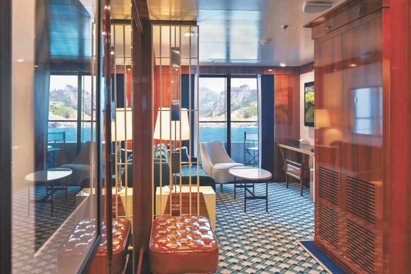 A view of the sitting area in the Navigator Suite on the 'World Traveller' with blue-and-white–patterned carpet, a leather bench, a sofa, an armchair, and a balcony