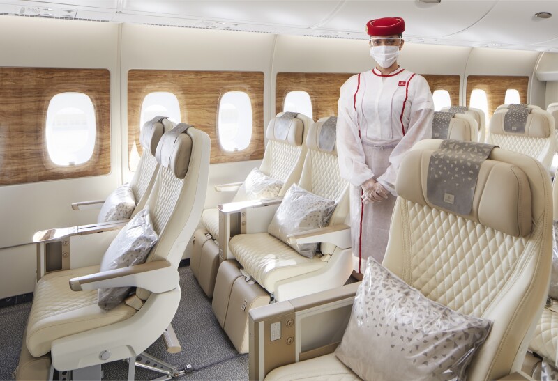 Empty white seats with silver pillows in Emirates' Premium Economy with a standing flight attendant, dressed in white with face mask and round red hat