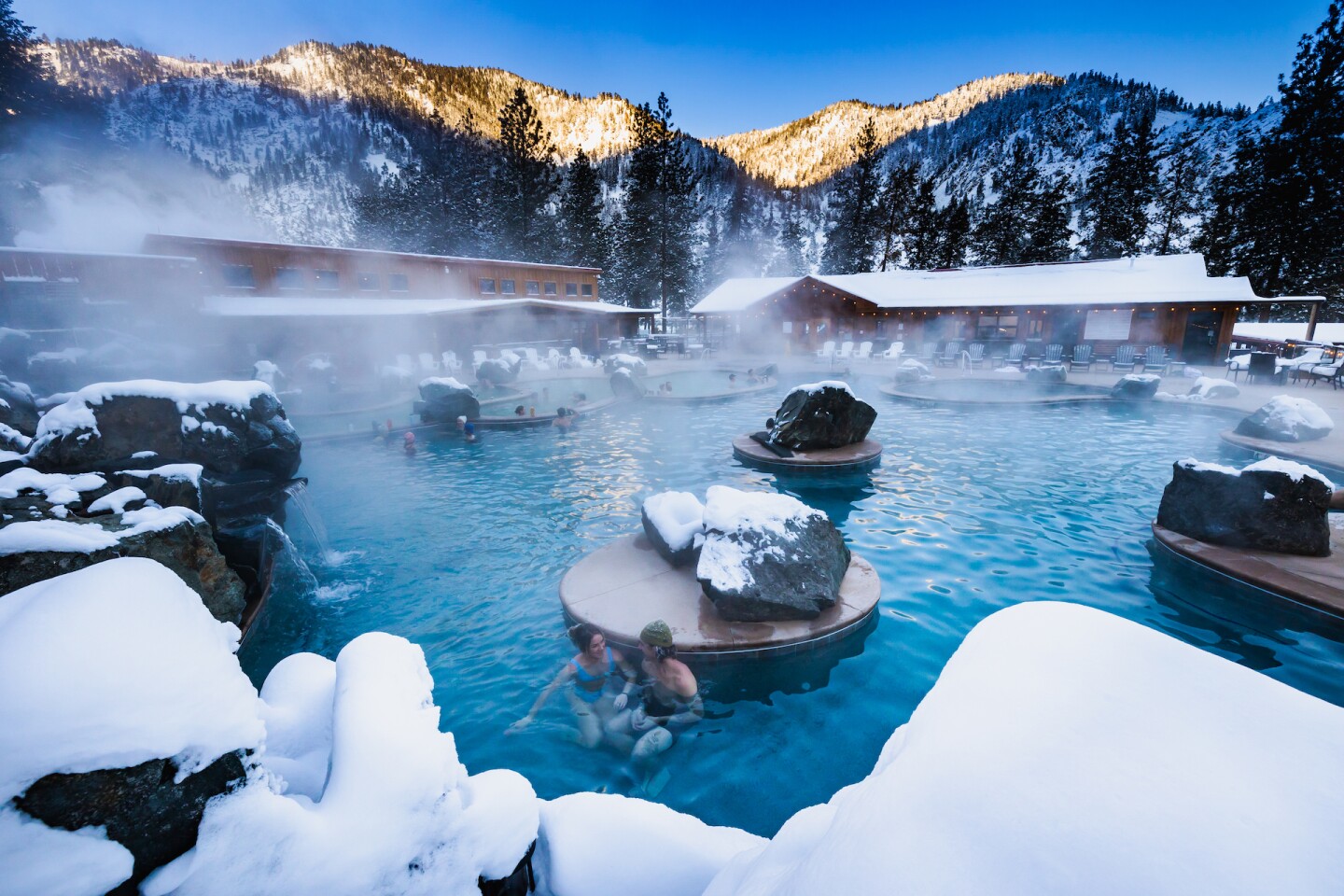 People bathing in the steaming hot springs at Quinn's Hot Springs Resort with snow in the foreground, and on the property roof and mountains in the background