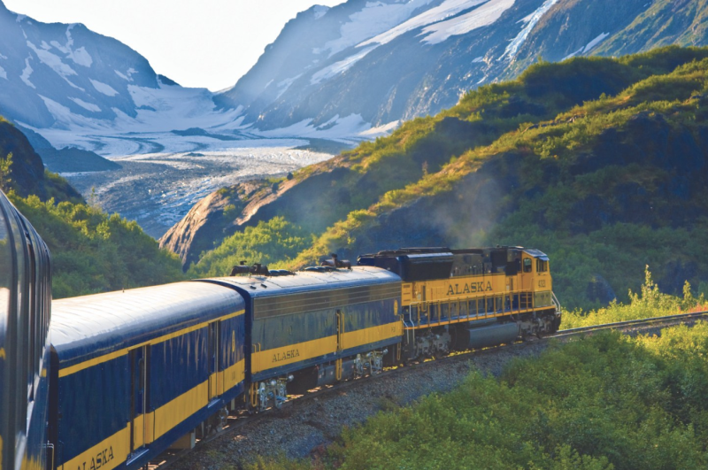 A blue-and-gold train curving around a bend, in front of a glacier.