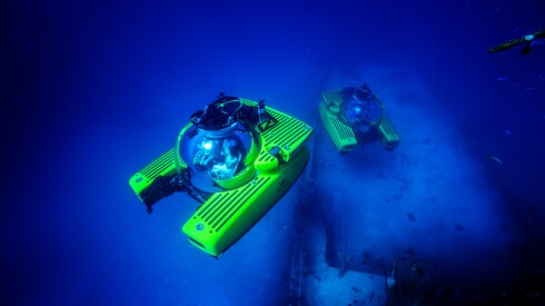 Two yellow Triton submarines underwater near a shipwreck