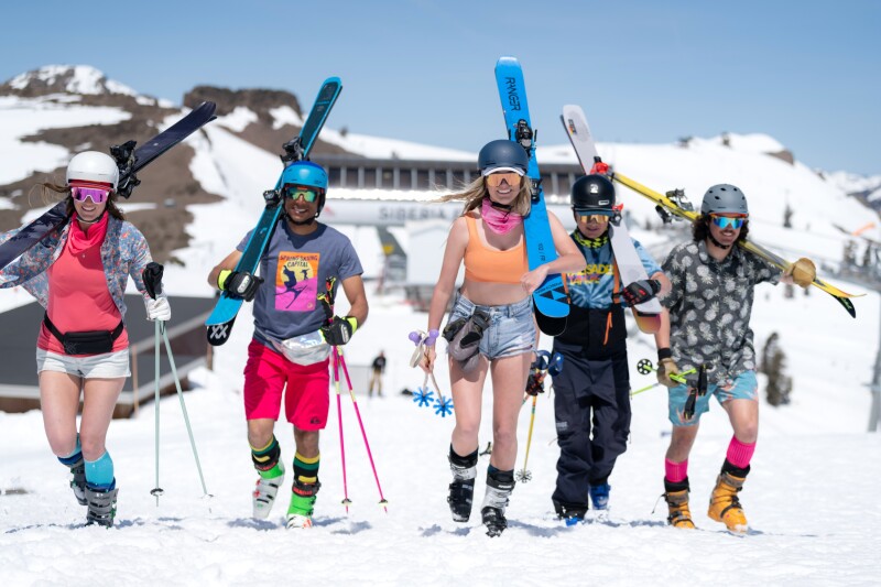 Five people carrying skis, dressed in colorful shorts, T-shirts, helmets, and snow boots, with snow on the ground