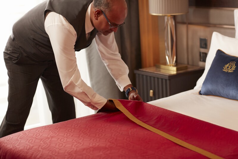 A Cunard butler making the bed in a cruise ship stateroom with a red duvet cover