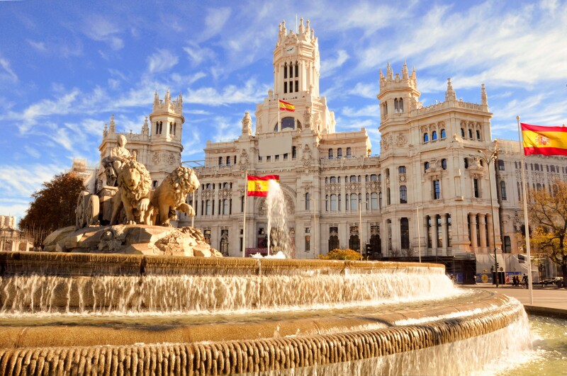 Large, circular Cibeles fountain in Madrid, with statue of lions in center and several red and yellow flags in background