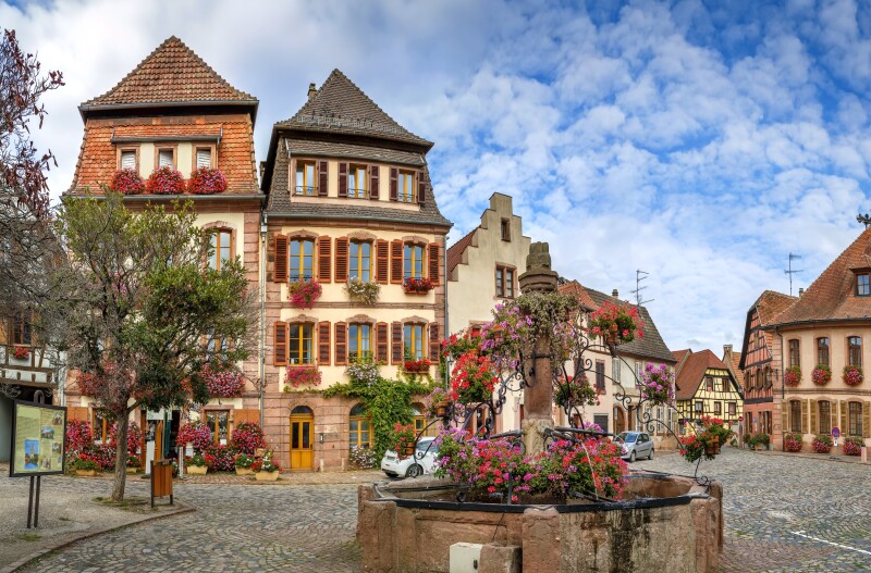 Half-timbered houses behind small round stone fountain with pots of red and purple flowers