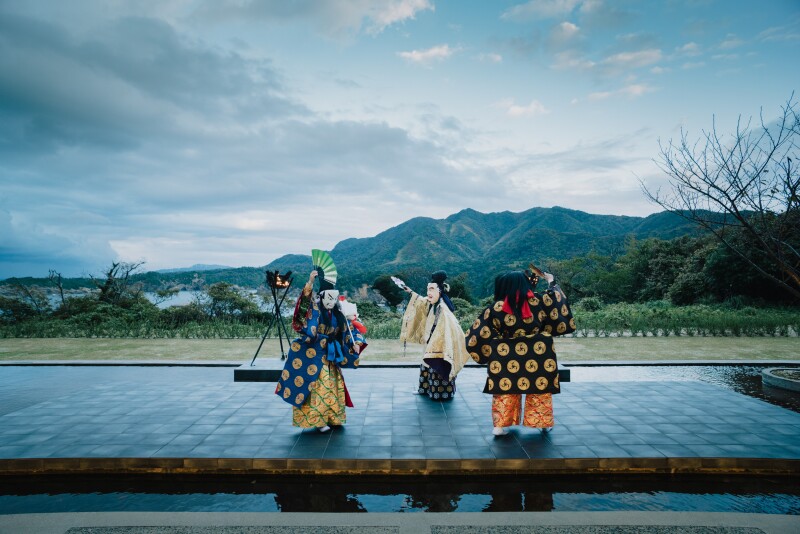 Three people in colorful costumes outdoors performing a ceremonial Shinto dance, with mountains in distance