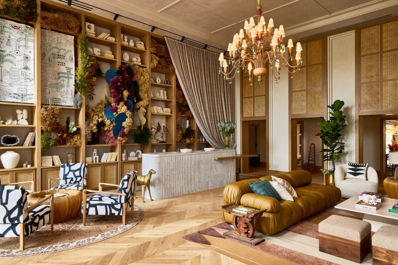 A lobby with low couches, patterned chairs, a sweeping curtain, chandelier, and a large floral installation creeping up a built-in bookcase