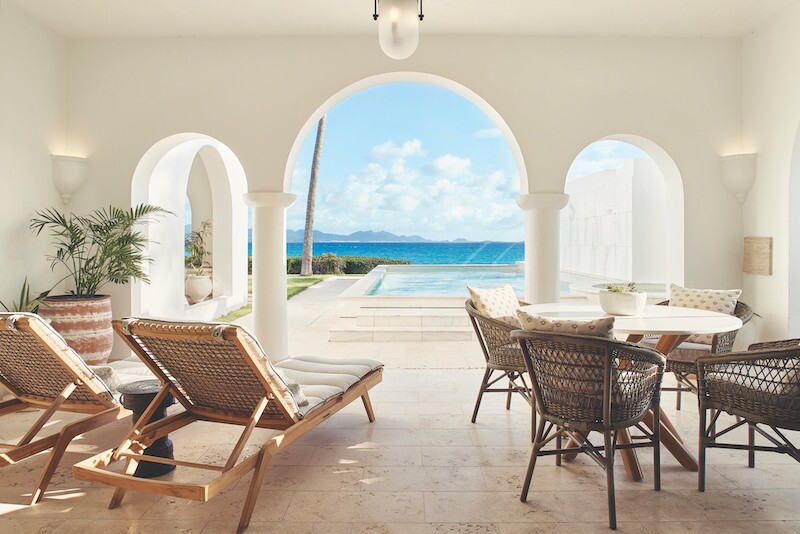 A few lounge chairs and table in white open-air room with three arched openings facing pool and sea at Cap Juluca