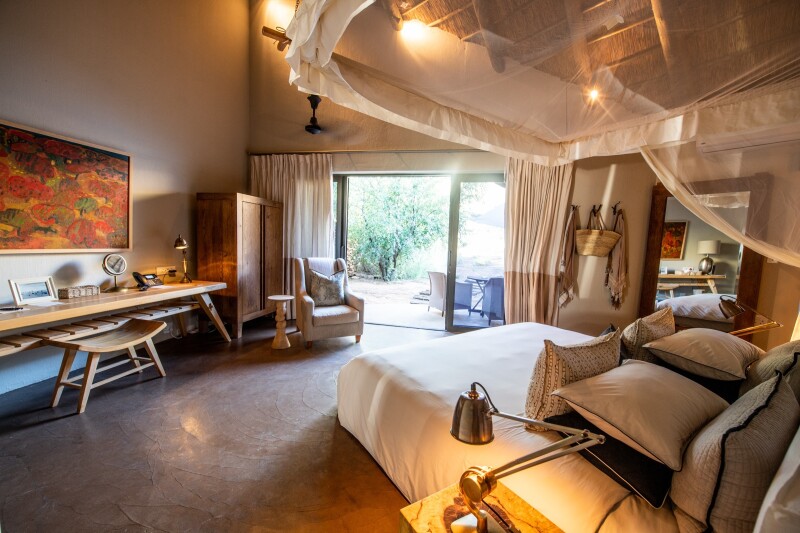 A large beige room at Tswalu Kalahari with bed topped by pillows and with net above, plus a desk and armchair, armoire, and outdoor patio area