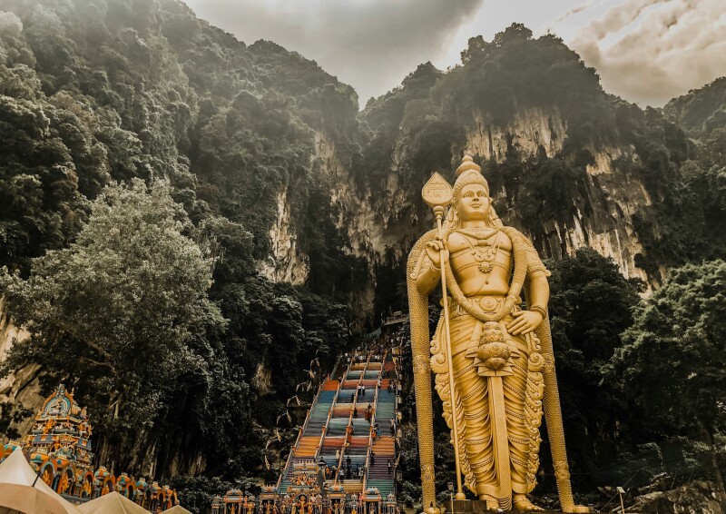 A massive gold statue flanks a series of painted steps that lead up to the Batu Caves, with craggy mountain covered in greenery in background