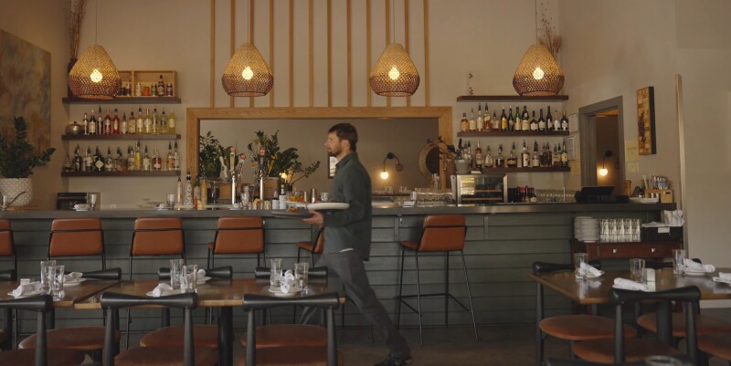 A waiter carries dishes across an empty bar and dining room. Warm earth tone colors accent the design of the Loma restaurant.