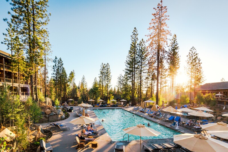 Large rectangular outdoor pool, with people lounging around it under beige umbrellas and tall pines in background