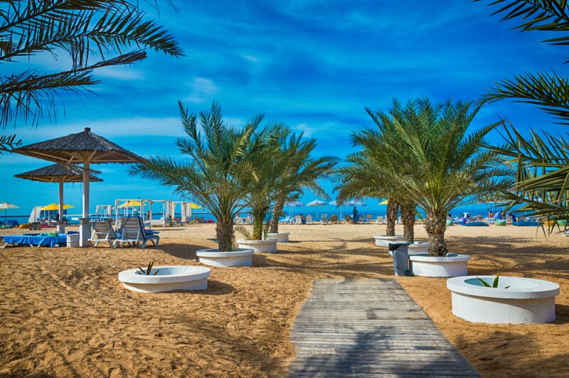 A golden-sand beach, with wooden boardwalk cutting through center, short palm trees in planters, and umbrellas