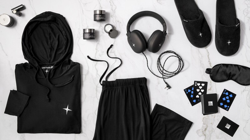 An overhead image of black pajamas with a hooded long-sleeve shirt, over-ear headphones, playing cards, black slippers, an eye mask, and Perricone MD beauty products