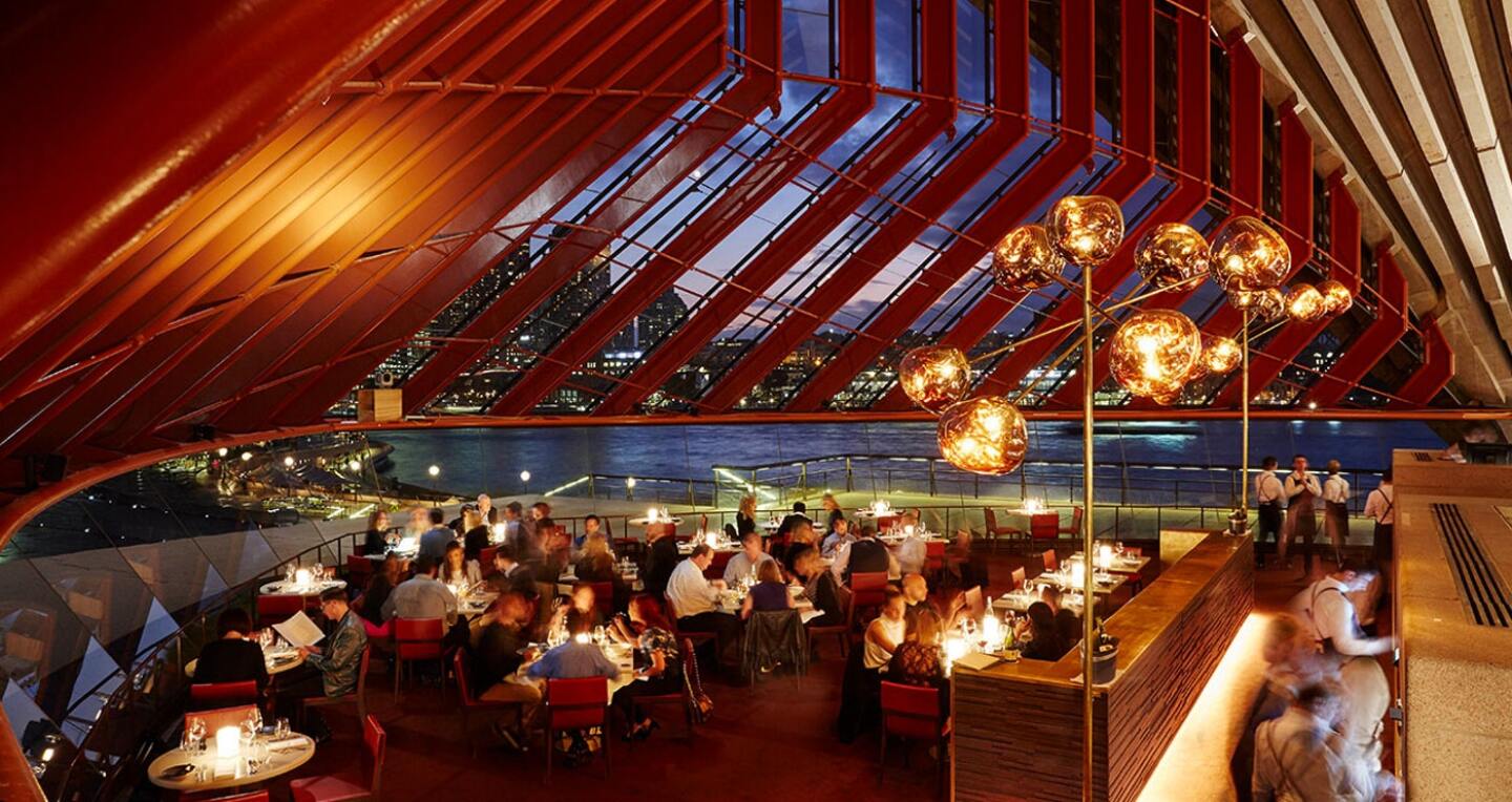 People sit around tables in a fancy restaurant. The evening skyline is seen through an angled glass roof.