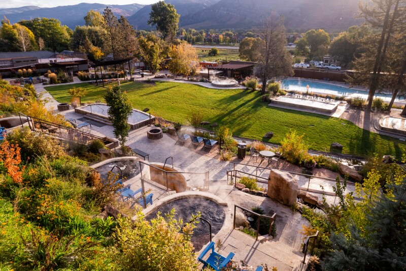 Aerial view of a complex of pools and hot tubs surrounded by green lawn and trees, with mountains in distance