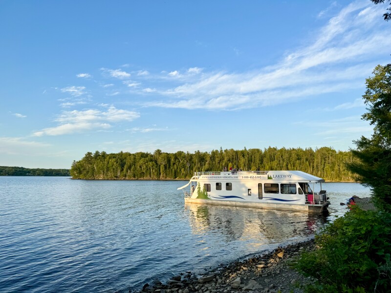 A white New Brunswick houseboat on calm river with shores lined by evergreens