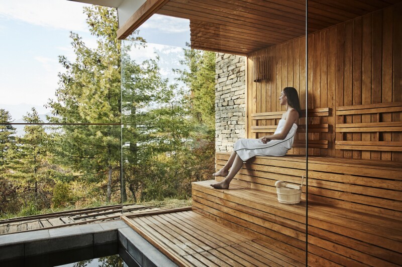 A woman wrapped in white towel sitting in sauna, with wall of windows overlooking trees