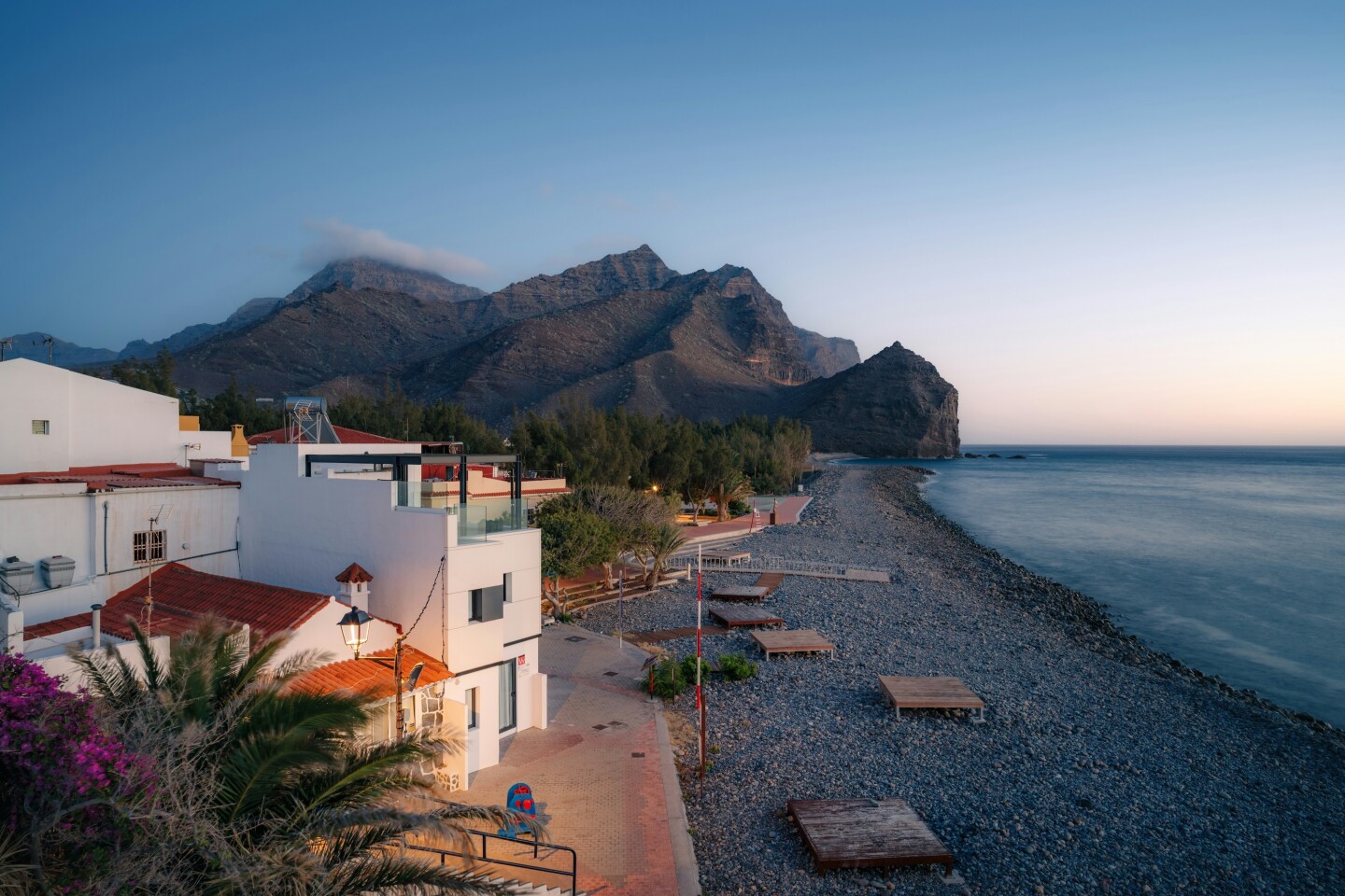 A white building on the beach at left, with mountains in the background and rocky beach with several flat, elevated lounge beds at right