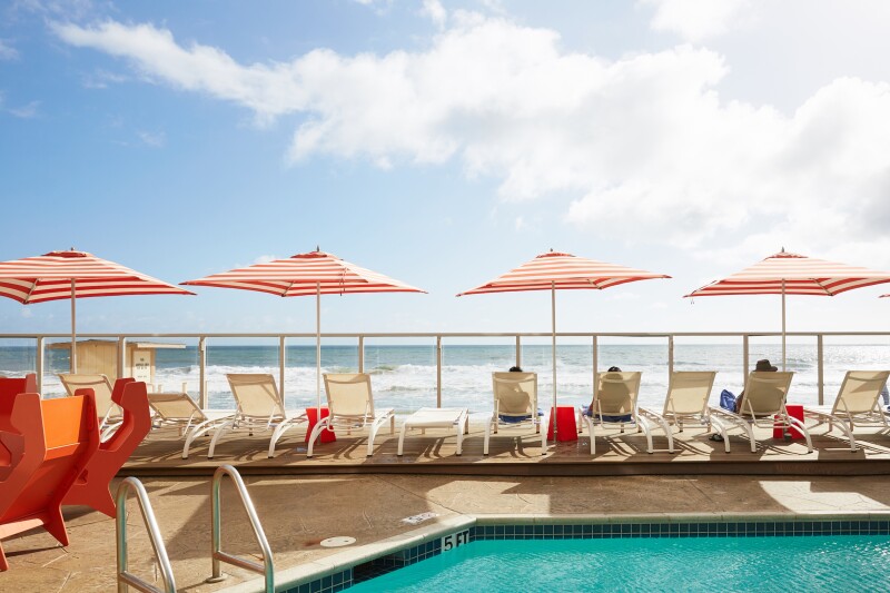 Row of pool chairs with a few people in them and four red-and-white striped sun umbrellas overlooking ocean