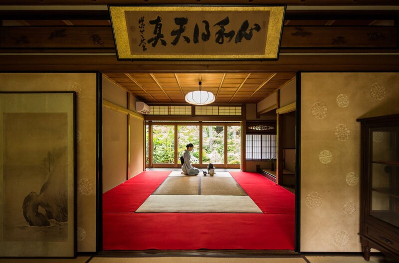 A traditionally dressed woman kneels in the center of a room before the items for a tea ceremony.