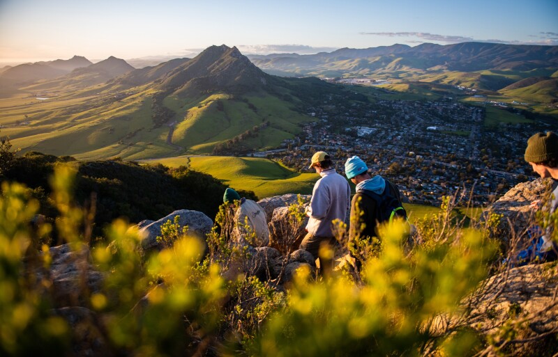 A group of hikers descend a mountain. Rolling green hills and the city of San Luis Obispo are below.