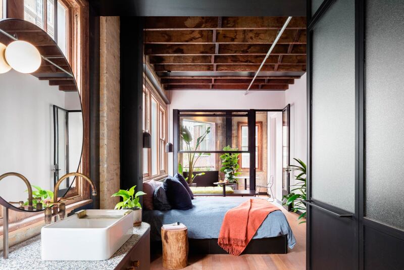 The bathroom leads into the bedroom. A gold faucet sits over a white, square sink with a circular mirror. The bed has a light blue comforter, dark blue pillows, and an orange throw blanket.