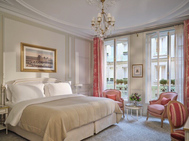 White guest room with small chandelier, pink wingback chairs, and mint green railing on balcony behind two large windows